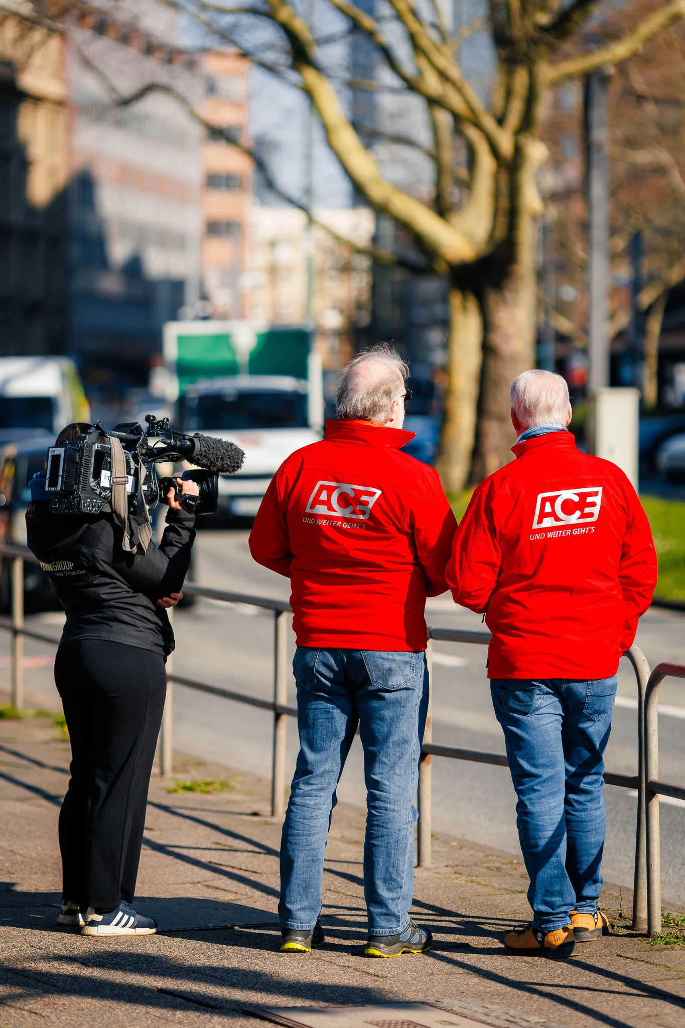 Zwei Ehrenamtliche in roten ACE Westen stehen an einer Straße mit einem Kameramann